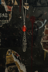 A gothic-style silver chain necklace featuring a faceted red gemstone and a dangling safety-pin style charm with a small cross, set against a dark, distressed graffiti wall.