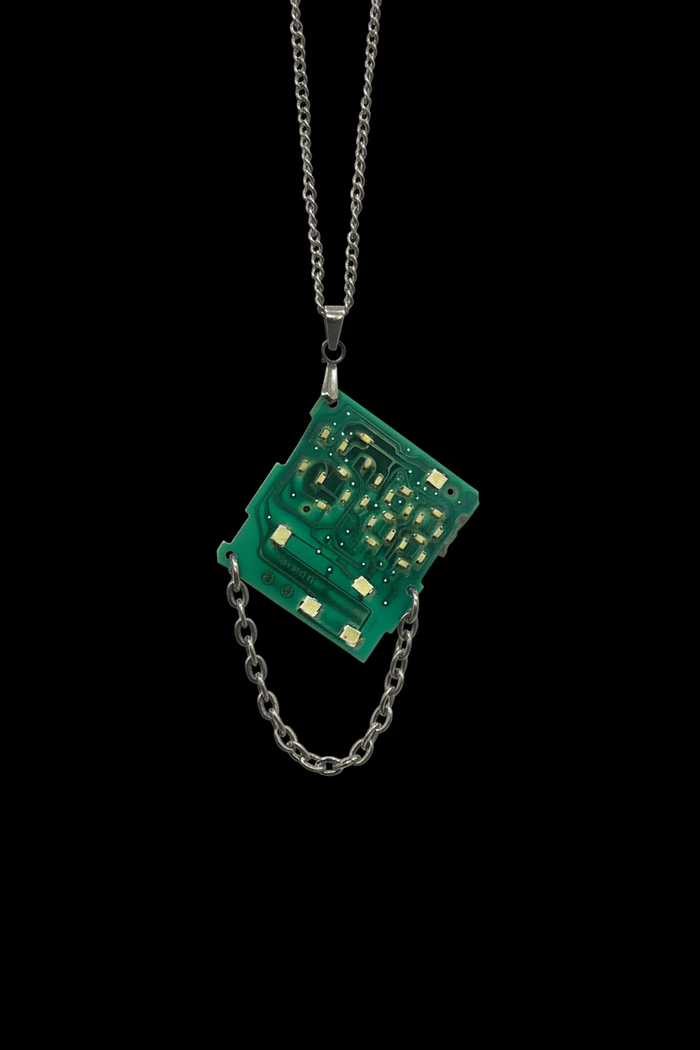 A green electronic circuit board pendant necklace on a silver chain, set against a solid black background.