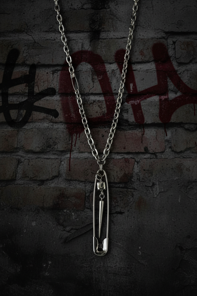 A silver safety-pin pendant necklace hanging against a dark brick wall with red graffiti.