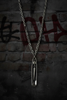 A silver safety-pin pendant necklace hanging against a dark brick wall with red graffiti.
